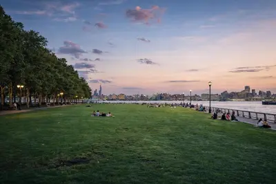 a group of people sitting on the grass near the water at Hoboken 15 Min Train to Manhattan Full Kitchen in Hoboken