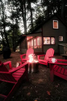 a group of red chairs around a fire pit in front of a house at Rustic, Pet Friendly Cabin on the Hudson River, Perfect for a Glamping Weekend in the Adirondacks in Corinth