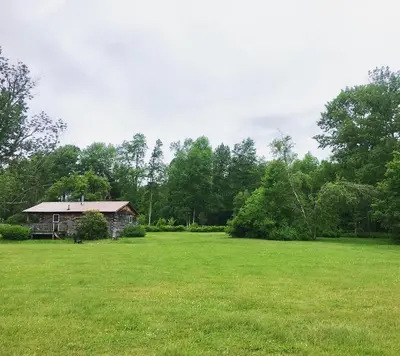 a house in a field of green grass with trees at Rustic Cabin Rental for Groups near Woodstock, Upstate New York in Big Indian