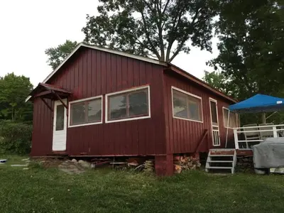 a red house is sitting in the grass at Cozy Cabin with Lovely Views of Great Sacandaga Lake in Northville, New York in Northville