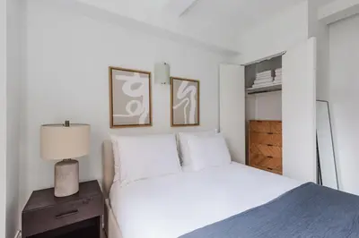 a white bedroom with a bed and a nightstand with a lamp at Blueground Upper West Side fitness center nr Central Park NYC-1596 in New York