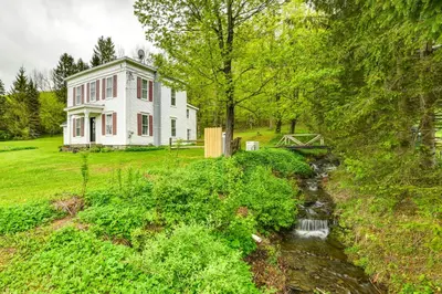 a white house with a creek in front of it at Hike and Baseball Near Cooperstown Home on 30 Acres in Westford