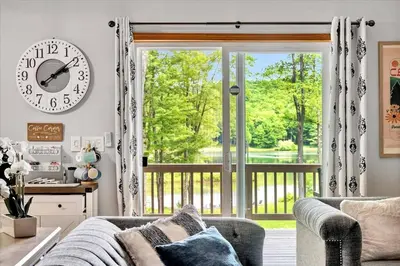 a living room with a large window and a clock at Wooded lakefront chalet with hot tub and game room in South Kortright