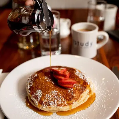 a person pouring syrup onto a plate of pancakes with strawberries at The Study at Yale, Study Hotels in New Haven
