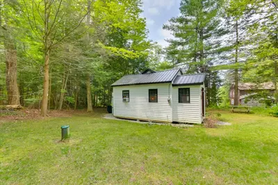 a white tiny house in a yard with trees at East Otis Vacation Rental with Private Deck and BBQ! in East Otis
