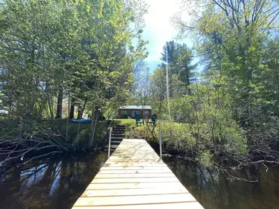 a wooden bridge over a river with a house in the background at Adirondack Waterfront Cabin Upper Hudson Tributary in Lake Luzerne