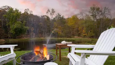 a fire pit next to two chairs and a lake at 4BDR Heritage Estate w Pool Private Pond Fire Pit Cortland by AvantStay in Saugerties